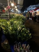 A bustling market scene at night featuring numerous bouquets of closed lilies arranged in plastic containers. The market is lit by overhead lights, while vendors and customers mill around in the background. Metal stalls and structures are visible, creating an industrial atmosphere.