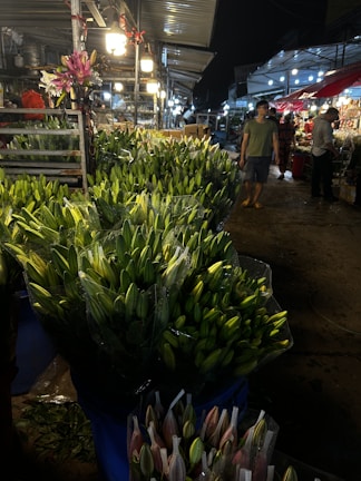 A bustling market scene at night featuring numerous bouquets of closed lilies arranged in plastic containers. The market is lit by overhead lights, while vendors and customers mill around in the background. Metal stalls and structures are visible, creating an industrial atmosphere.