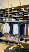A clothing store display featuring neatly folded sweatshirts and t-shirts on wooden shelves, along with a range of hanging garments such as hoodies and shirts. The foreground shows a table with neatly folded pants and an empty wooden hanger.