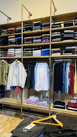 A clothing store display featuring neatly folded sweatshirts and t-shirts on wooden shelves, along with a range of hanging garments such as hoodies and shirts. The foreground shows a table with neatly folded pants and an empty wooden hanger.