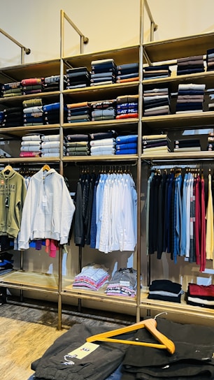 A clothing store display featuring neatly folded sweatshirts and t-shirts on wooden shelves, along with a range of hanging garments such as hoodies and shirts. The foreground shows a table with neatly folded pants and an empty wooden hanger.