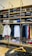 A clothing store display featuring neatly folded sweatshirts and t-shirts on wooden shelves, along with a range of hanging garments such as hoodies and shirts. The foreground shows a table with neatly folded pants and an empty wooden hanger.