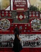 A vibrant mosaic wall with predominantly red tiles and various colorful patterns and artworks is on display. The phrase 'Brasil, eu te amo Selaron' is prominently featured in white lettering against a red background. A woman with long dark hair, wearing a black dress, stands in front of the mosaic, looking at the artwork. The wall includes several intricate designs and colorful tiling, portraying boats and cityscapes in different sections.