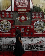 A vibrant mosaic wall with predominantly red tiles and various colorful patterns and artworks is on display. The phrase 'Brasil, eu te amo Selaron' is prominently featured in white lettering against a red background. A woman with long dark hair, wearing a black dress, stands in front of the mosaic, looking at the artwork. The wall includes several intricate designs and colorful tiling, portraying boats and cityscapes in different sections.