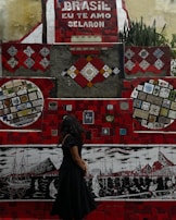 A vibrant mosaic wall with predominantly red tiles and various colorful patterns and artworks is on display. The phrase 'Brasil, eu te amo Selaron' is prominently featured in white lettering against a red background. A woman with long dark hair, wearing a black dress, stands in front of the mosaic, looking at the artwork. The wall includes several intricate designs and colorful tiling, portraying boats and cityscapes in different sections.