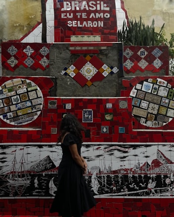 A vibrant mosaic wall with predominantly red tiles and various colorful patterns and artworks is on display. The phrase 'Brasil, eu te amo Selaron' is prominently featured in white lettering against a red background. A woman with long dark hair, wearing a black dress, stands in front of the mosaic, looking at the artwork. The wall includes several intricate designs and colorful tiling, portraying boats and cityscapes in different sections.