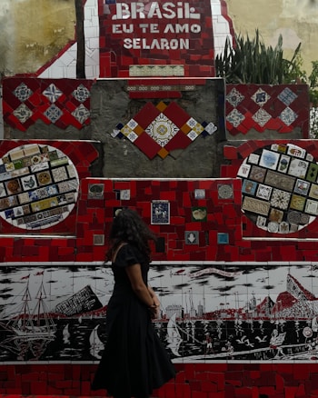 A vibrant mosaic wall with predominantly red tiles and various colorful patterns and artworks is on display. The phrase 'Brasil, eu te amo Selaron' is prominently featured in white lettering against a red background. A woman with long dark hair, wearing a black dress, stands in front of the mosaic, looking at the artwork. The wall includes several intricate designs and colorful tiling, portraying boats and cityscapes in different sections.