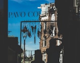 A clothesline with various lingerie items is silhouetted against a bright blue sky. Two figures can be seen in silhouette, one wearing a hat and the other with their hand on their head. The background reveals part of a brick building and a streetlamp, with the words 'Pavo Couture Amsterdam' visible on the window glass.