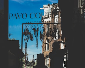 A clothesline with various lingerie items is silhouetted against a bright blue sky. Two figures can be seen in silhouette, one wearing a hat and the other with their hand on their head. The background reveals part of a brick building and a streetlamp, with the words 'Pavo Couture Amsterdam' visible on the window glass.
