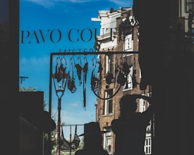 A clothesline with various lingerie items is silhouetted against a bright blue sky. Two figures can be seen in silhouette, one wearing a hat and the other with their hand on their head. The background reveals part of a brick building and a streetlamp, with the words 'Pavo Couture Amsterdam' visible on the window glass.