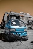 A damaged blue van with a broken front bumper is parked in front of a building with large signs. The building has signage indicating a bakery and auto express shop. The sky is a sepia tone, creating a contrast with the vivid color of the van.