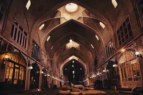 The image depicts an indoor marketplace with an arched ceiling. The architecture is characterized by intricate brickwork and numerous arched windows and lamps hanging from the ceiling. The space appears to be dimly lit with a warm, golden hue radiating from the lamps. There are several stacks of goods or rugs laid out on the floor in the middle of the walkway.