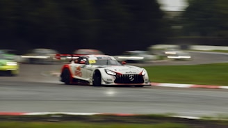 A racing car with a distinctive white and red color scheme speeds through a wet race track, creating motion blur. The car's sleek design features prominent branding and a large rear spoiler. Other cars are visible in the background, adding to the sense of a competitive race in progress. The surrounding area includes green grass and blurred spectators.