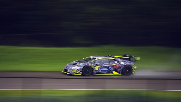 A sleek, high-performance racing car is speeding along a blurred racetrack, indicating significant motion. The car features a vibrant yellow and purple livery with various sponsor logos and a prominent rear spoiler.