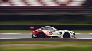 A sleek racing car speeds along a track, captured in motion with a blurred background. The car has a white body with red and black accents, and noticeable branding and sponsor logos. The car's aerodynamic design and large rear spoiler suggest high performance.