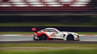 A sleek racing car speeds along a track, captured in motion with a blurred background. The car has a white body with red and black accents, and noticeable branding and sponsor logos. The car's aerodynamic design and large rear spoiler suggest high performance.