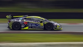 A sleek, high-performance sports car is captured in motion on a racetrack. The vehicle features a dynamic, colorful design with geometric patterns and logos. Bright neon yellow accents highlight its aerodynamic shape, contrasting with its dark body paint. Blurred background elements emphasize the intense speed of the car.