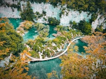 A scenic landscape featuring a winding wooden walkway over turquoise waters surrounded by lush greenery and rocky cliffs. The water appears to be flowing gently, creating a serene and peaceful atmosphere. Trees with vibrant leaves frame the image, adding a touch of autumnal orange to the scene.
