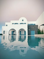 A modern Mediterranean-style building features whitewashed walls and blue doors and windows. The structure has multiple arches and is reflected in a tranquil swimming pool in the foreground. A stone wall segment adds texture to the facade, complemented by sparse greenery.