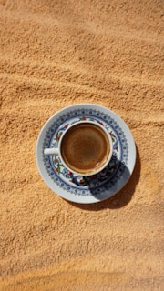 A intricately designed cup of espresso with a decorative floral pattern sits on a matching saucer against a textured sandy background. The coffee appears slightly frothy.