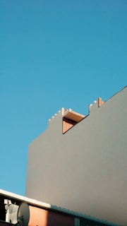 A minimalist urban scene featuring a portion of a building with a simple light gray facade against a clear blue sky. The building has distinctive geometric shapes and a visible brick structure near the top. A section of guttering and a satellite dish can be seen in the lower left corner.