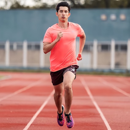 A person is running on a red track in an outdoor setting. They are wearing a bright pink athletic shirt, black shorts, and pink running shoes with black accents. The background shows a blue wall and blurred elements, indicating depth of field.