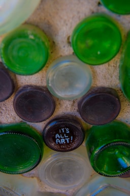 A close-up view of an arrangement of bottle bottoms embedded in a cement-like surface. Some are green while others are clear, and one of the brown ones has the words 'IT'S ALL ART!' written on it.