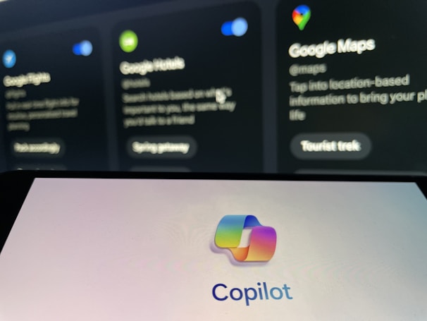 The image shows a blurred background with displays of Google services such as Google Flights, Google Hotels, and Google Maps. In the foreground, there is a clear view of a screen displaying the Copilot logo with colorful overlapping shapes.
