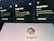 The image shows a blurred background with displays of Google services such as Google Flights, Google Hotels, and Google Maps. In the foreground, there is a clear view of a screen displaying the Copilot logo with colorful overlapping shapes.
