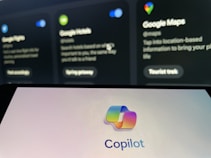 The image shows a blurred background with displays of Google services such as Google Flights, Google Hotels, and Google Maps. In the foreground, there is a clear view of a screen displaying the Copilot logo with colorful overlapping shapes.