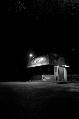 A dark street scene featuring a small closed shop or booth illuminated by a dim street light. The environment is desolate with minimal visibility due to the surrounding darkness. The mood is emphasized by the black and white color scheme.