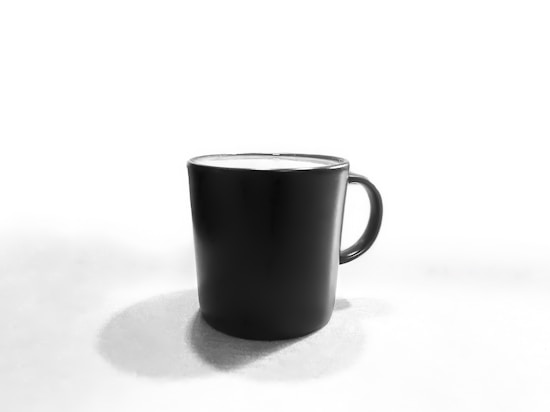 A black ceramic mug stands against a stark white background. The mug casts a soft shadow on the surface it rests on, indicating a light source from above. Its glossy finish reflects ambient light, creating a subtle contrast against the matte background.