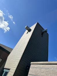 A tall, modern building structure made of light-colored bricks extends upwards towards a bright blue sky with a few scattered clouds. The building features two large external lights attached high on the wall.