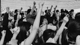 A large group of people with raised fists, signifying unity or protest. The image is in black and white and captures diverse expressions of determination and solidarity. Some individuals are wearing masks and the crowd is dense.