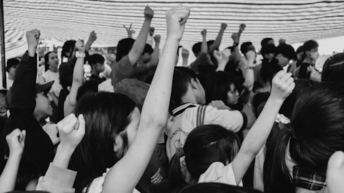 A large group of people with raised fists, signifying unity or protest. The image is in black and white and captures diverse expressions of determination and solidarity. Some individuals are wearing masks and the crowd is dense.