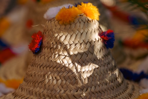 A woven straw hat decorated with colorful pom-poms in shades of red, orange, and yellow. The sunlight creates a play of light and shadows on the textured surface, highlighting the intricate weaving pattern.