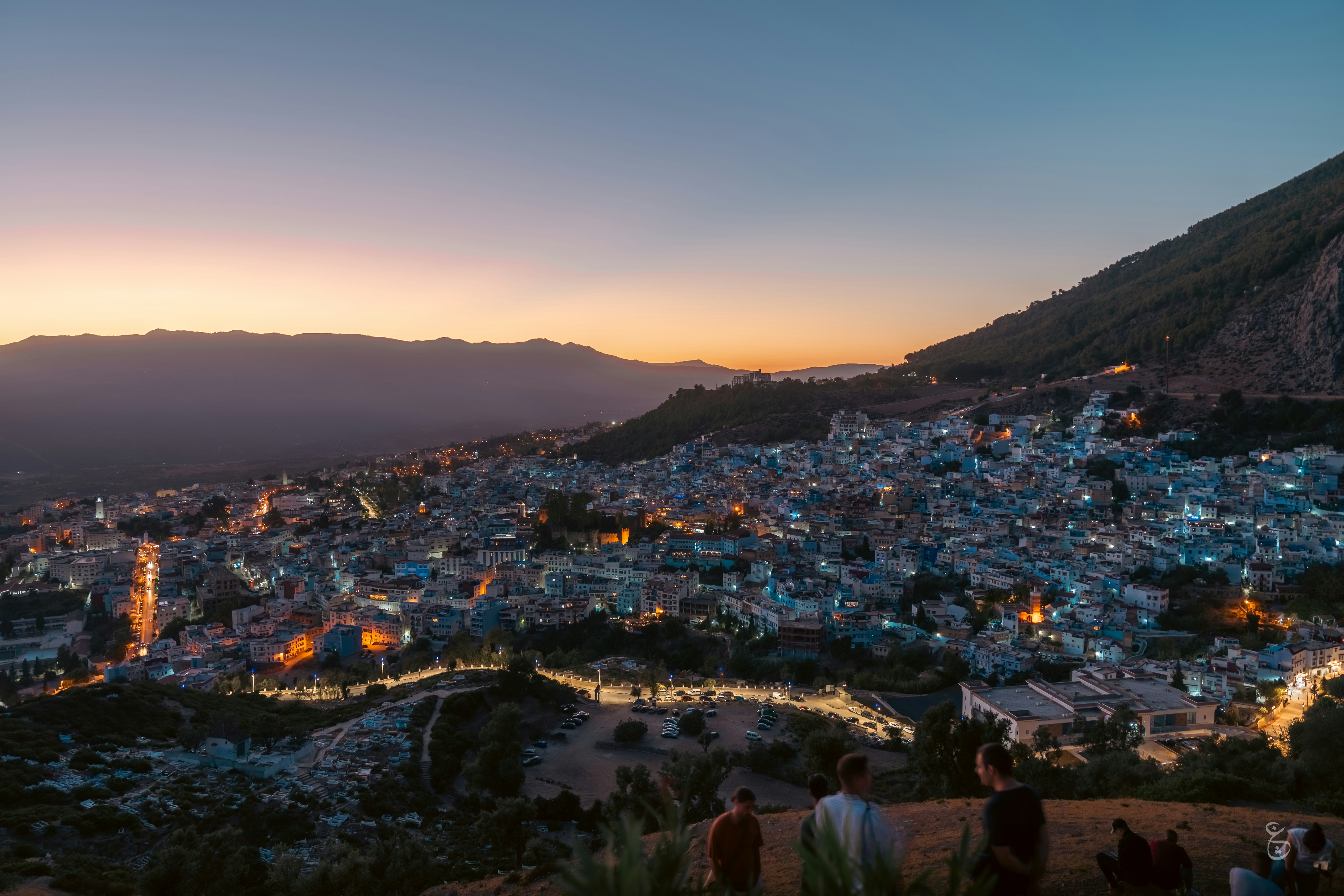 a group of people standing on top of a hill, the blue city moments after the sun sets behind the mountains.