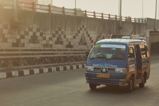 A blue minibus with the branding 'Batik Solo Trans' is driving on a road next to a barrier wall featuring a geometric black and white pattern. The atmosphere appears to be slightly hazy, indicating possible early morning or late afternoon sunlight.