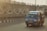 A blue minibus with the branding 'Batik Solo Trans' is driving on a road next to a barrier wall featuring a geometric black and white pattern. The atmosphere appears to be slightly hazy, indicating possible early morning or late afternoon sunlight.