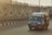 A blue minibus with the branding 'Batik Solo Trans' is driving on a road next to a barrier wall featuring a geometric black and white pattern. The atmosphere appears to be slightly hazy, indicating possible early morning or late afternoon sunlight.