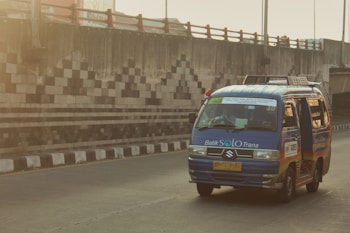 A blue minibus with the branding 'Batik Solo Trans' is driving on a road next to a barrier wall featuring a geometric black and white pattern. The atmosphere appears to be slightly hazy, indicating possible early morning or late afternoon sunlight.
