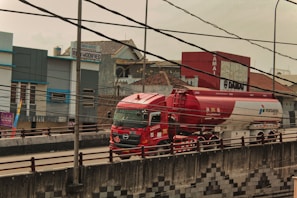 A red and white fuel tanker truck is driving on a multi-lane road with buildings in the background. There are numerous electrical cables overhead, crossing in multiple directions. The truck has several logos and text on its body, and the road has a concrete barrier with a pattern design. The surrounding area includes commercial buildings and signs.