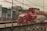 A red and white fuel tanker truck is driving on a multi-lane road with buildings in the background. There are numerous electrical cables overhead, crossing in multiple directions. The truck has several logos and text on its body, and the road has a concrete barrier with a pattern design. The surrounding area includes commercial buildings and signs.