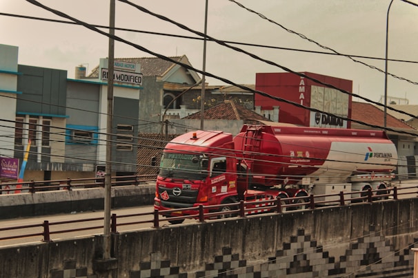 A red and white fuel tanker truck is driving on a multi-lane road with buildings in the background. There are numerous electrical cables overhead, crossing in multiple directions. The truck has several logos and text on its body, and the road has a concrete barrier with a pattern design. The surrounding area includes commercial buildings and signs.