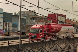A red and white fuel tanker truck is driving on a multi-lane road with buildings in the background. There are numerous electrical cables overhead, crossing in multiple directions. The truck has several logos and text on its body, and the road has a concrete barrier with a pattern design. The surrounding area includes commercial buildings and signs.