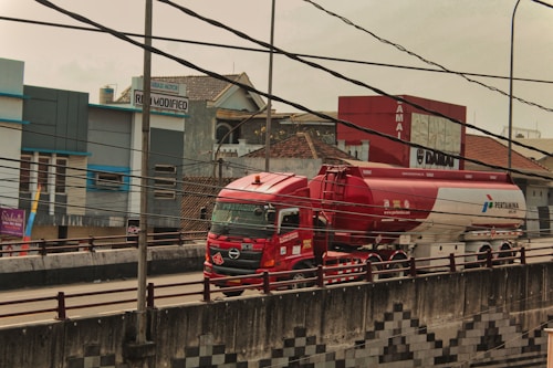 A red and white fuel tanker truck is driving on a multi-lane road with buildings in the background. There are numerous electrical cables overhead, crossing in multiple directions. The truck has several logos and text on its body, and the road has a concrete barrier with a pattern design. The surrounding area includes commercial buildings and signs.