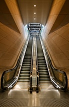 Two parallel escalators ascend within a modern architectural structure featuring warm brown paneling. The ceiling lights above illuminate the metal and glass surfaces, leading the eye towards an overhead framework that suggests a higher level or exit.