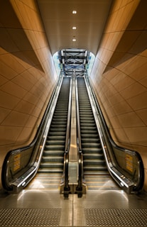 Two parallel escalators ascend within a modern architectural structure featuring warm brown paneling. The ceiling lights above illuminate the metal and glass surfaces, leading the eye towards an overhead framework that suggests a higher level or exit.