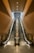 Two parallel escalators ascend within a modern architectural structure featuring warm brown paneling. The ceiling lights above illuminate the metal and glass surfaces, leading the eye towards an overhead framework that suggests a higher level or exit.