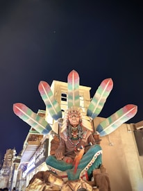 A statue of a Native American figure sitting cross-legged, adorned with a headdress featuring large colorful feathers. The background is a clear night sky, and the scene is brightly lit from below, highlighting the ornate attire of the statue.